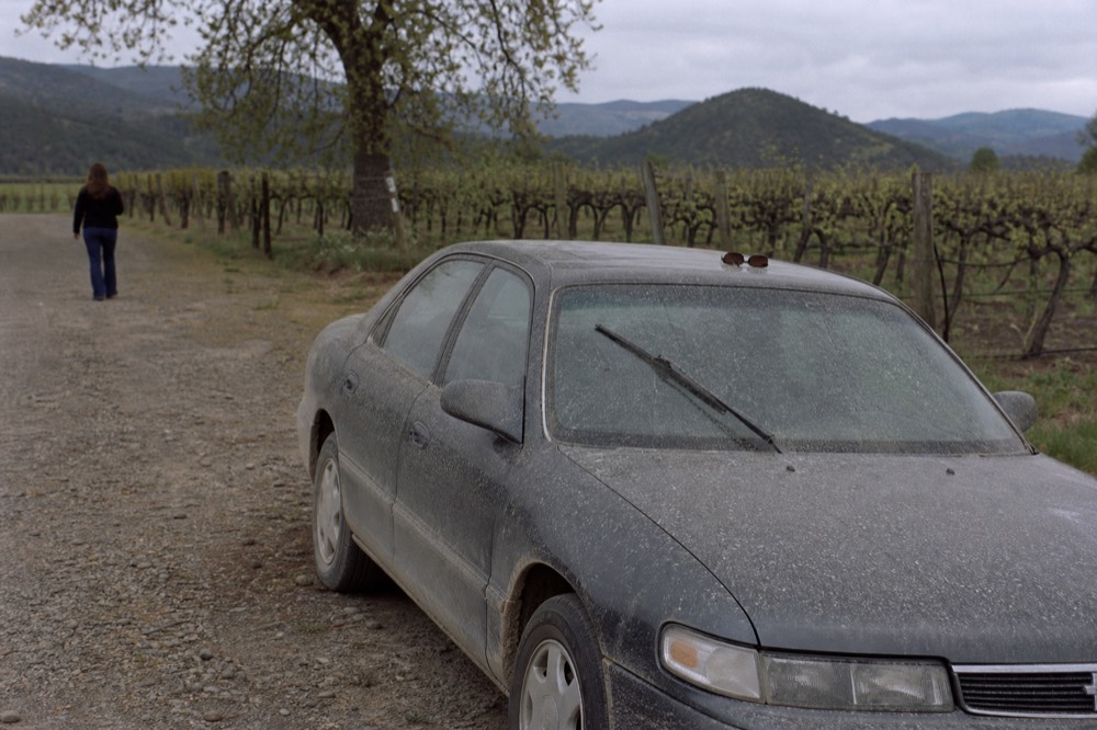 A parked car beside a gravel winery driveway with hills behind, no text, no logos, no people.