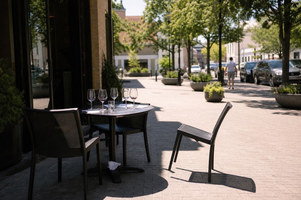 A sidewalk table with wine glasses outside a tasting room on a town plaza, no text, no logos, no people.